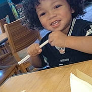 Renall is registered to the contest to win money with this photo: child, toddler, curly_hair, smiling, indoor, restaurant, table, wooden_chair, straw, black_shirt, playing, happy, face, person, young, casual, background, arcade_machine, furniture, light