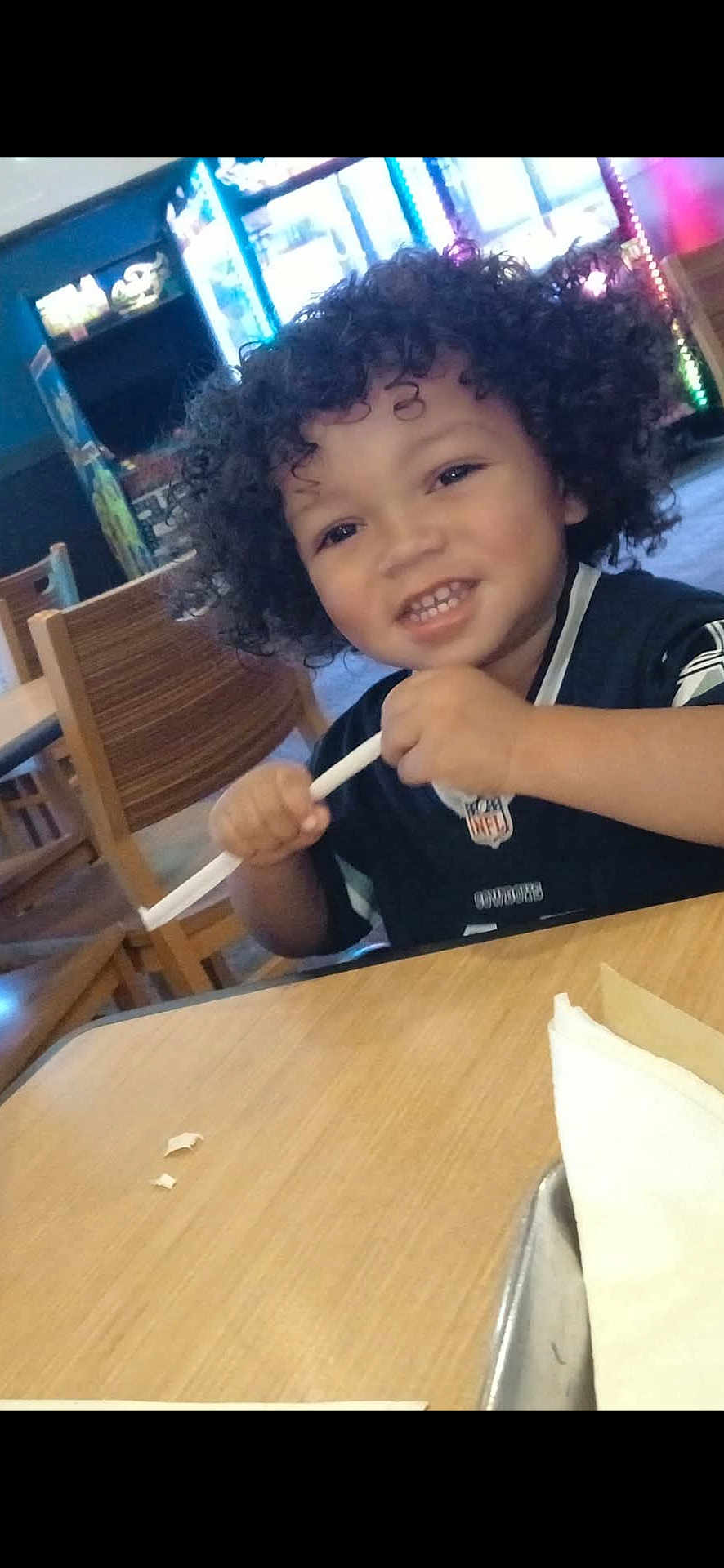 Renall is registered to the contest to win money with this photo: child, toddler, curly_hair, smiling, indoor, restaurant, table, wooden_chair, straw, black_shirt, playing, happy, face, person, young, casual, background, arcade_machine, furniture, light