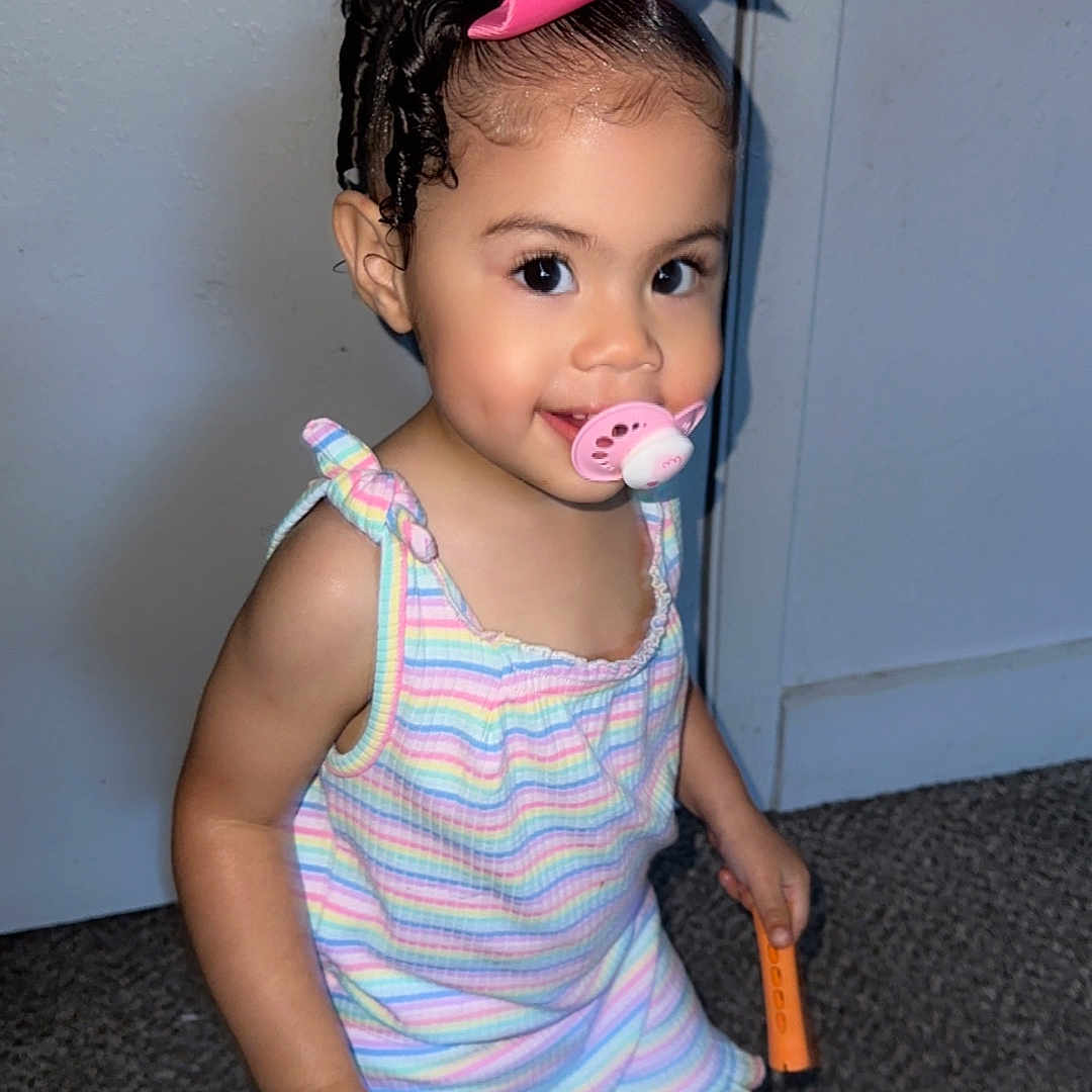 Zion joined the competition — help win amazing prizes! bow, braided_hair, carpet, child, cute, door, fashion, happy, holding, indoor, pacifier, person, portrait, romper, shoes, smile, striped_clothing, toddler, toy, young_child