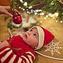 baby, child, christmas, christmas_ornament, christmas_tree, cute, festive, floor, hand, holiday, holiday_decoration, indoors, looking_up, red_clothing, santa_hat, snowflake, striped_sleeves, tree_skirt, twinkling_lights, wooden_floor