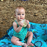 Maysie is registered to the contest to win money with this photo: baby, child, swimsuit, towel, rocky_ground, messy, cream, face, hands, legs, blue, turquoise, sitting, outdoor, cute, small_cup, curious, person, summer, playful