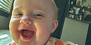 Maysie is registered to the contest to win money with this photo: baby, child, smiling, close_up, drooling, indoor, natural_light, window, face, head, person, happy, expression, colorful_clothing, patterned_clothing, room, furniture, shelf, door, portrait