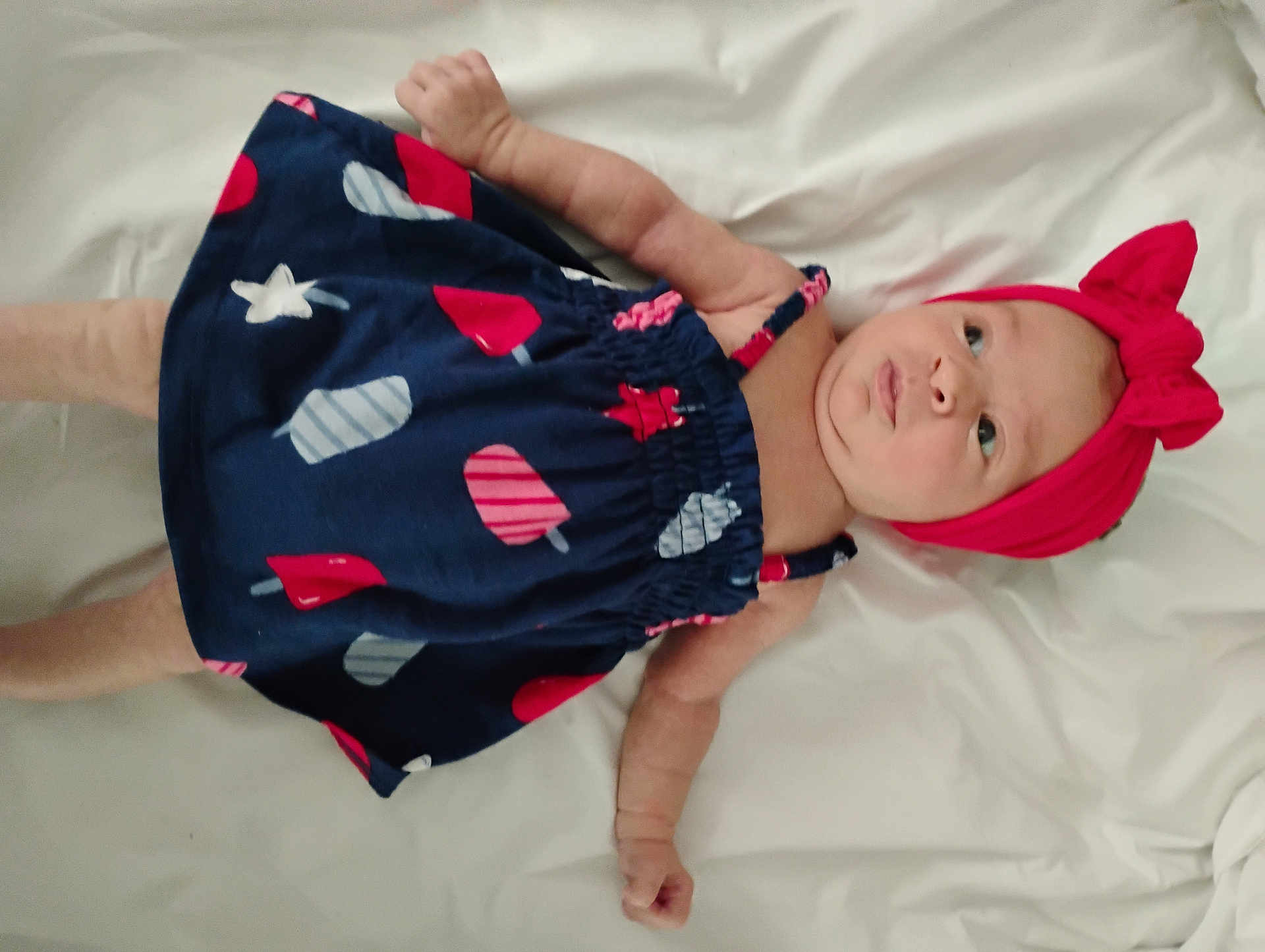 Addilyn is registered to the contest to win money with this photo: baby, infant, child, dress, headband, bow, navy_blue, red, popsicle_print, lying_down, white_background, cute, soft, skin, arms, legs, clothing, portrait, indoors, curious