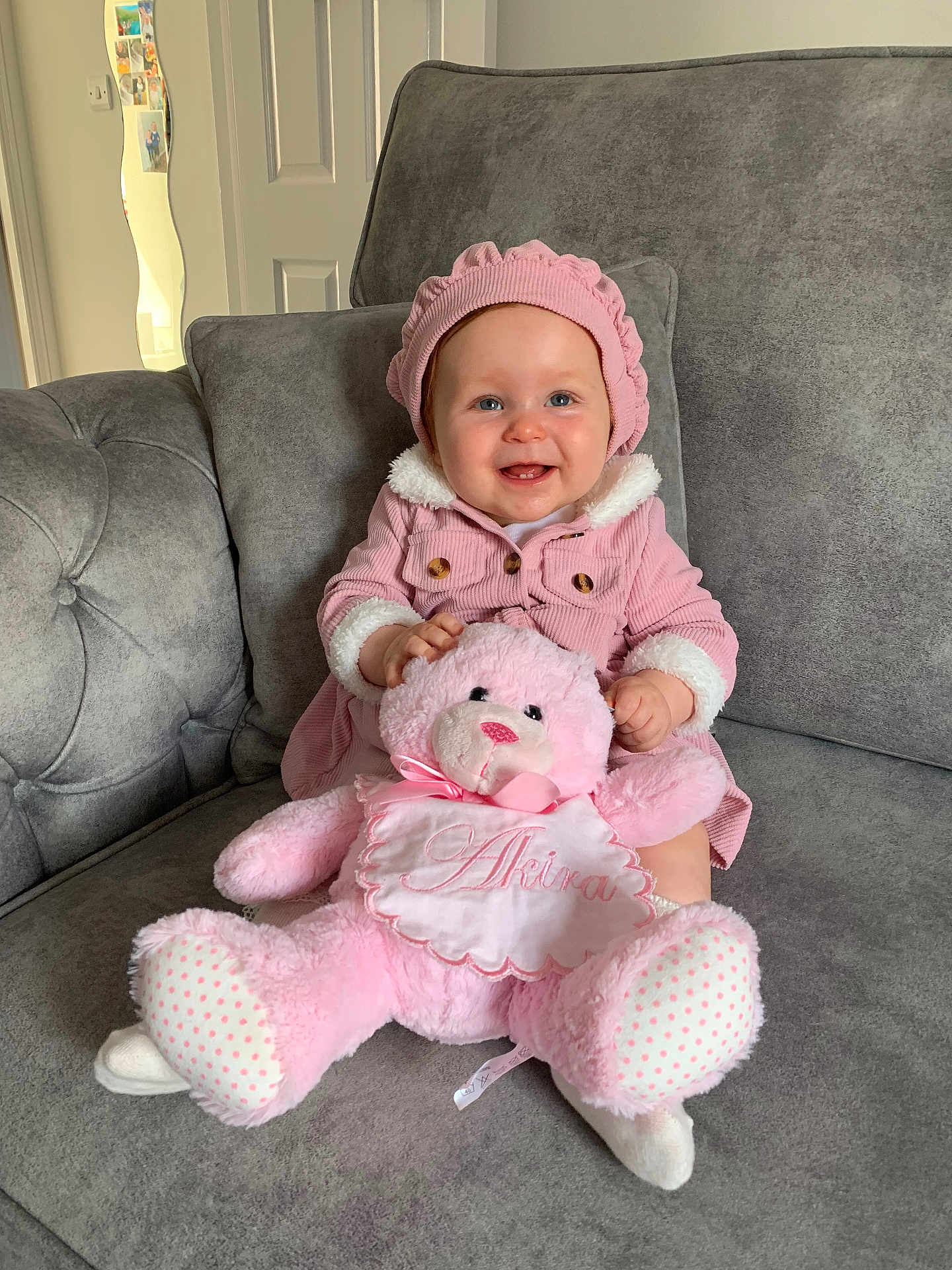 Akira is registered to the contest to win money with this photo: baby, child, pink_clothing, bonnet, teddy_bear, plush_toy, couch, grey_furniture, smiling, happy, indoors, portrait, cute, soft_toy, seated, cozy, infant, home, warm_lighting, comfort