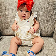 Cataleya is registered to the contest to win money with this photo: baby, child, red_bow, dress, blue_sandals, couch, seated, curious_expression, indoors, cute, infant, footwear, holding_clothing, plush_furniture, portrait, headwear, bright_colors, skin, legs, comfortable