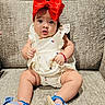 baby, child, red_bow, dress, blue_sandals, couch, seated, curious_expression, indoors, cute, infant, footwear, holding_clothing, plush_furniture, portrait, headwear, bright_colors, skin, legs, comfortable