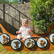 Björn a rejoint le concours — aidez-le/la à gagner de superbes lots ! baby, child, viking_hat, wooden_ship, outdoor, grass, sunlight, shield, letters, smiling, greenery, stone_wall, costume, playful, portrait, sitting, summer, daylight, hat, cute