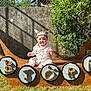 baby, child, viking_hat, wooden_ship, outdoor, grass, sunlight, shield, letters, smiling, greenery, stone_wall, costume, playful, portrait, sitting, summer, daylight, hat, cute