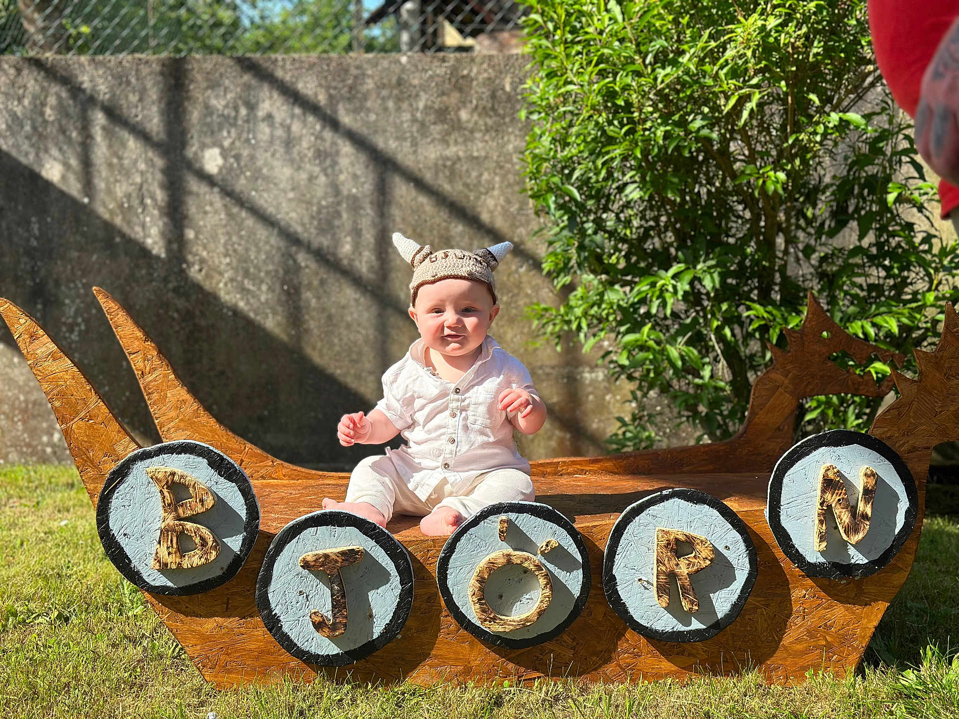 Björn a rejoint le concours — aidez-le/la à gagner de superbes lots ! baby, child, viking_hat, wooden_ship, outdoor, grass, sunlight, shield, letters, smiling, greenery, stone_wall, costume, playful, portrait, sitting, summer, daylight, hat, cute