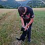 man, dog, field, grass, mountains, outdoor, nature, pet, playing, black_dog, white_markings, casual_clothing, shoes, smiling, happy, sunset, scenic, landscape, summer, fun