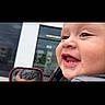 baby, infant, child, smiling, gummy_smile, face, portrait, close_up, car, vehicle, side_mirror, outdoor, daylight, building, window, reflection, gray_clothing, happy, candid, photography