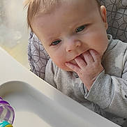 Kinslee is registered to the contest to win money with this photo: baby, infant, highchair, teething, fingers_in_mouth, toy, plastic_toy, window_light, sunlight, patterned_cushion, onesie, hand, face, cheeks, hair, tray, portrait, indoor, curious, sitting