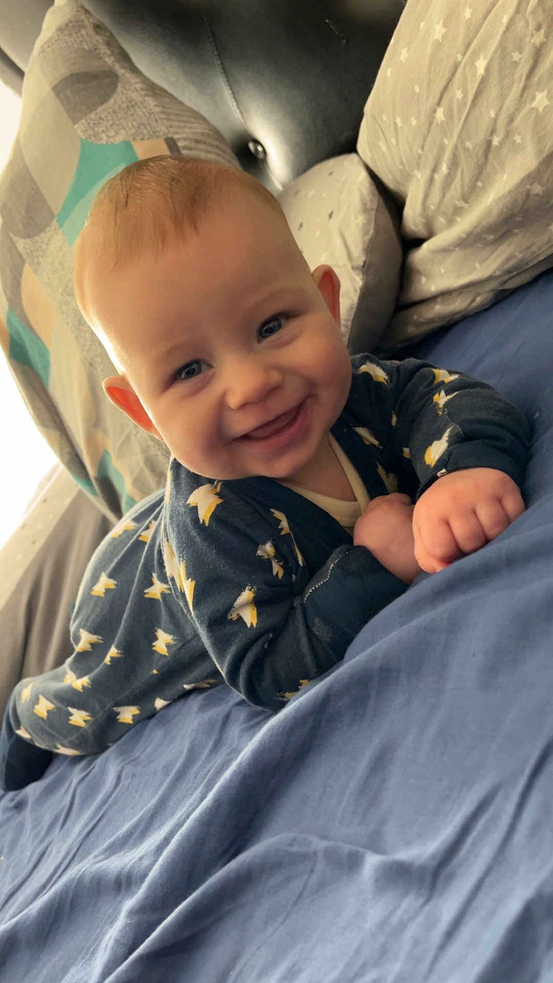 Edzyo participe au concours pour gagner de l'argent avec cette photo : baby, infant, smiling, onesie, bedding, pillow, blanket, blue_bedspread, stars_pattern, lying_down, close_up, face, eyes, hands, tummy_time, indoor, portrait, happy, hair, cheek