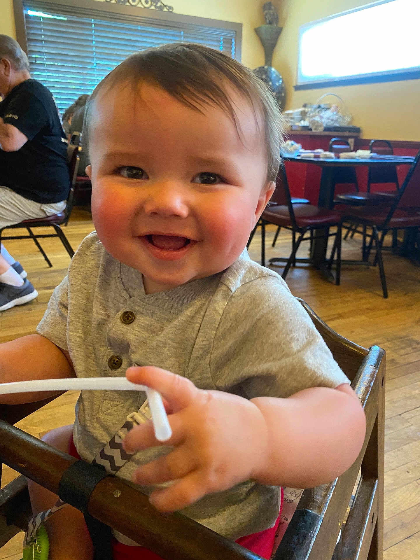 River is registered to the contest to win money with this photo: baby, child, smiling, high_chair, indoor, wooden_floor, restaurant, table, chair, person, happy, cute, short_hair, casual_clothing, holding, straw, playful, seated, daylight, cheerful