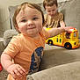 toddler, child, indoor, toy, bus, couch, playing, smiling, person, short_hair, orange_shirt, brown_shirt, furniture, carpet, window_blinds, happy, cute, baby, home, floor