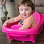 child, toddler, pink_toy, plastic, indoor, living_room, couch, floor, toy_ball, smiling, face, person, hair, clothing, seat, playful, curious, young_child, furniture, happy