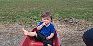 Weston is registered to the contest to win money with this photo: toddler, child, wagon, red_wagon, outdoor, grass, lake, trees, sky, clouds, snack, blue_shirt, black_shorts, barefoot, dirt, rocks, playing, nature, summer, park