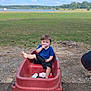 toddler, child, wagon, red_wagon, outdoor, grass, lake, trees, sky, clouds, snack, blue_shirt, black_shorts, barefoot, dirt, rocks, playing, nature, summer, park