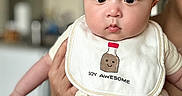 Oliver is registered to the contest to win money with this photo: apparel, baby, bib, child, chubby, clothing, cute, expression, eyes, face, funny_text, head, holding, human, indoors, infant, person, skin, smile, young