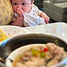 baby, infant, thumb_sucking, cloth, cushion, yellow, face, eyes, indoor, table, food, blurred_background, person, seated, curious, skin, head, hand, expression, closeup