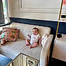 baby, baby_sitting, bib, cabinet, child, clothing, couch, face, floor, furniture, home_decor, indoor, pillow, reflection, smiling, stuffed_animal, table, toy, wall, window