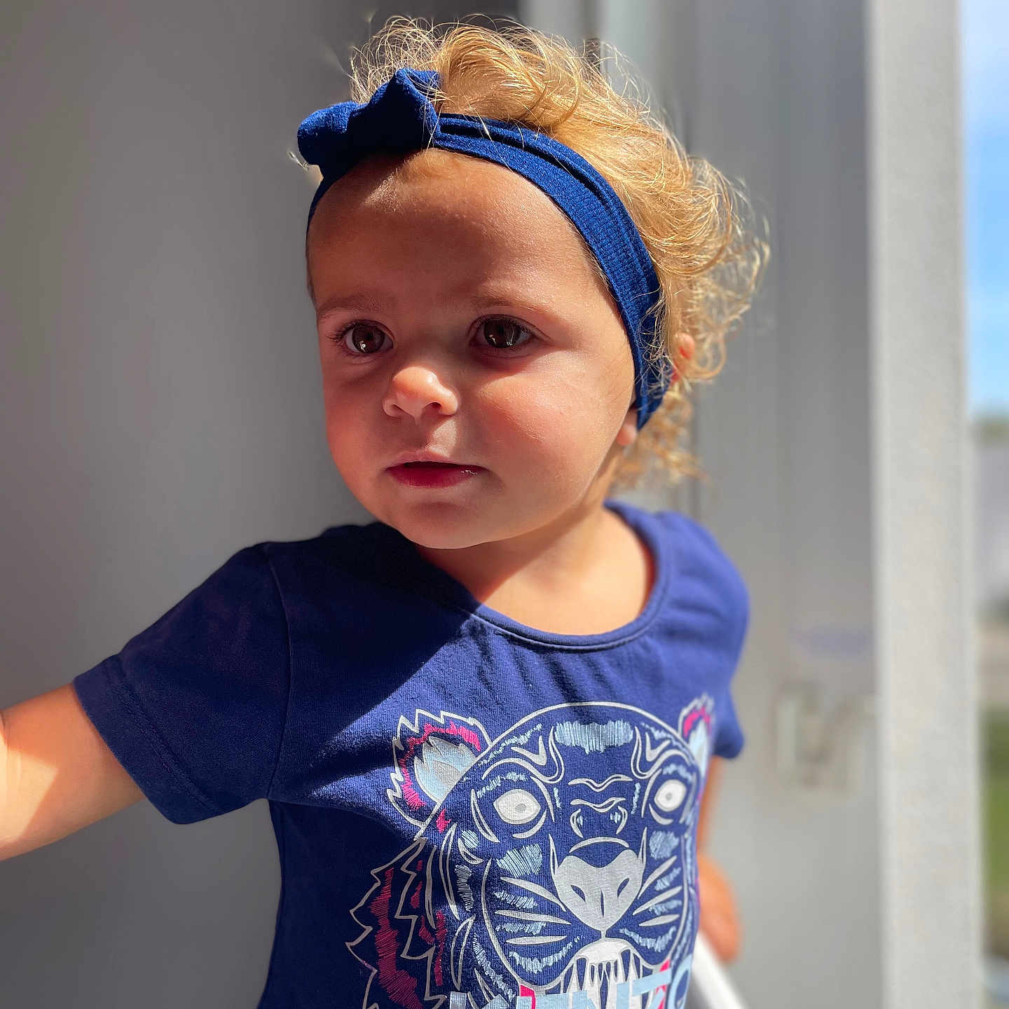 Yenna participe au concours pour gagner de l'argent avec cette photo : blue_shirt, casual_clothing, child, curly_hair, cute, daylight, expression, face, headband, indoor, looking_away, natural_light, person, portrait, short_sleeves, standing, tiger_graphic, toddler, window_light, young_child