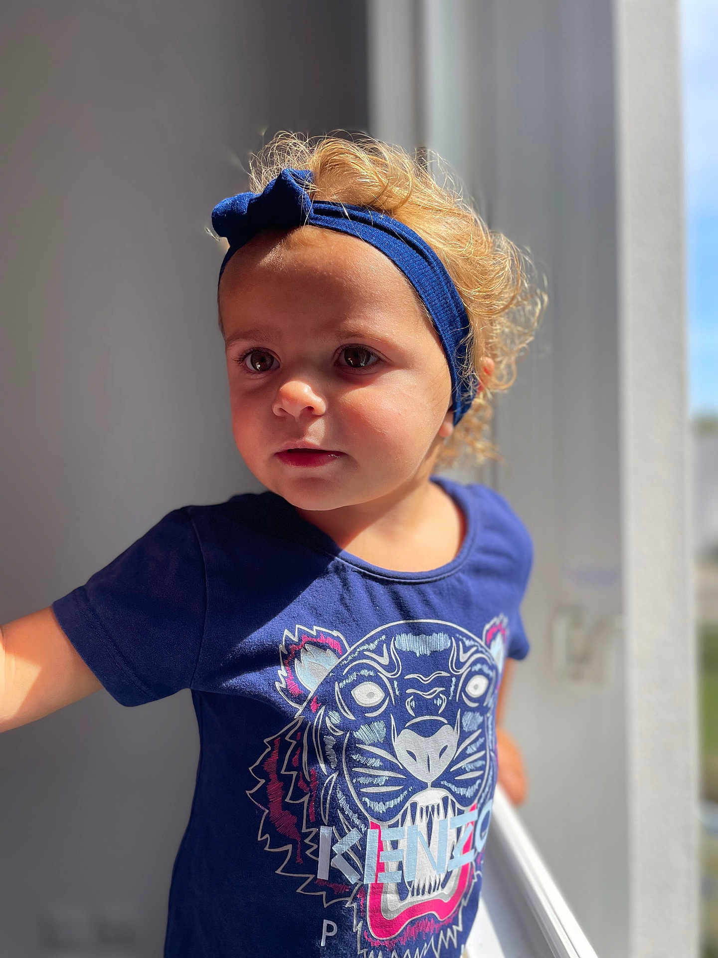 Yenna participe au concours pour gagner de l'argent avec cette photo : blue_shirt, casual_clothing, child, curly_hair, cute, daylight, expression, face, headband, indoor, looking_away, natural_light, person, portrait, short_sleeves, standing, tiger_graphic, toddler, window_light, young_child