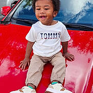Ace is registered to the contest to win money with this photo: child, toddler, boy, smiling, outdoor, car, red_car, hood, sitting, portrait, afro_hair, casual_clothing, white_shirt, pants, sneakers, fashion, parked_cars, trees, fence, daylight