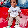child, toddler, boy, smiling, outdoor, car, red_car, hood, sitting, portrait, afro_hair, casual_clothing, white_shirt, pants, sneakers, fashion, parked_cars, trees, fence, daylight
