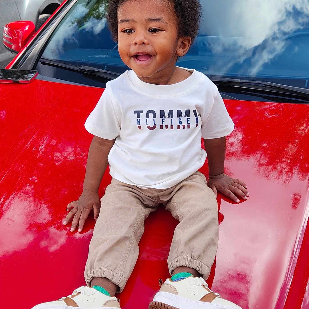 Ace is registered to the contest to win money with this photo: child, toddler, boy, smiling, outdoor, car, red_car, hood, sitting, portrait, afro_hair, casual_clothing, white_shirt, pants, sneakers, fashion, parked_cars, trees, fence, daylight