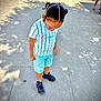 child, boy, standing, pavement, outdoor, daylight, shadows, braided_hair, striped_shirt, shorts, sneakers, looking_down, person, casual_clothing, sunlight, sidewalk, street, urban, summer, footwear