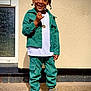 child, smiling, standing, green_clothing, corduroy, boots, necklace, outdoor, wall, concrete, fashion, happy, portrait, person, casual, sunlight, braids, pants, jacket, footwear