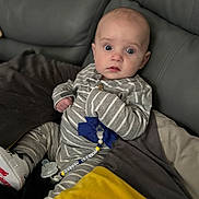 Jayden is registered to the contest to win money with this photo: baby, blanket, blue_bow, closeup, couch, curious_expression, face, footwear, hand, indoor, infant, pacifier, pajamas, person, pillow, seated, soft_furniture, striped_clothing, white_shoes, young_child