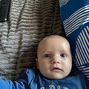 Jayden is registered to the contest to win money with this photo: baby, child, infant, blue_shirt, text_on_clothing, blanket, striped_pillow, bed, face, eyes, person, indoor, soft_texture, cute, closeup, expression, lying_down, young_child, head, small_hand