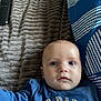 baby, child, infant, blue_shirt, text_on_clothing, blanket, striped_pillow, bed, face, eyes, person, indoor, soft_texture, cute, closeup, expression, lying_down, young_child, head, small_hand
