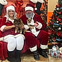 woman, man, dog, christmas_tree, holiday_decor, stockings, santa_claus, mrs_claus, glasses, white_hair, beard, red_clothing, white_trim, boots, tiled_floor, gift_box, plush_toy, smile, indoor, festive