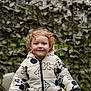 child, toddler, curly_hair, puffer_jacket, mickey_pattern, outdoor, garden, toy_vehicle, smile, portrait, winter_clothing, fence, leafy_background, grass, face, blue_eyes, standing, hands, zipper, cute