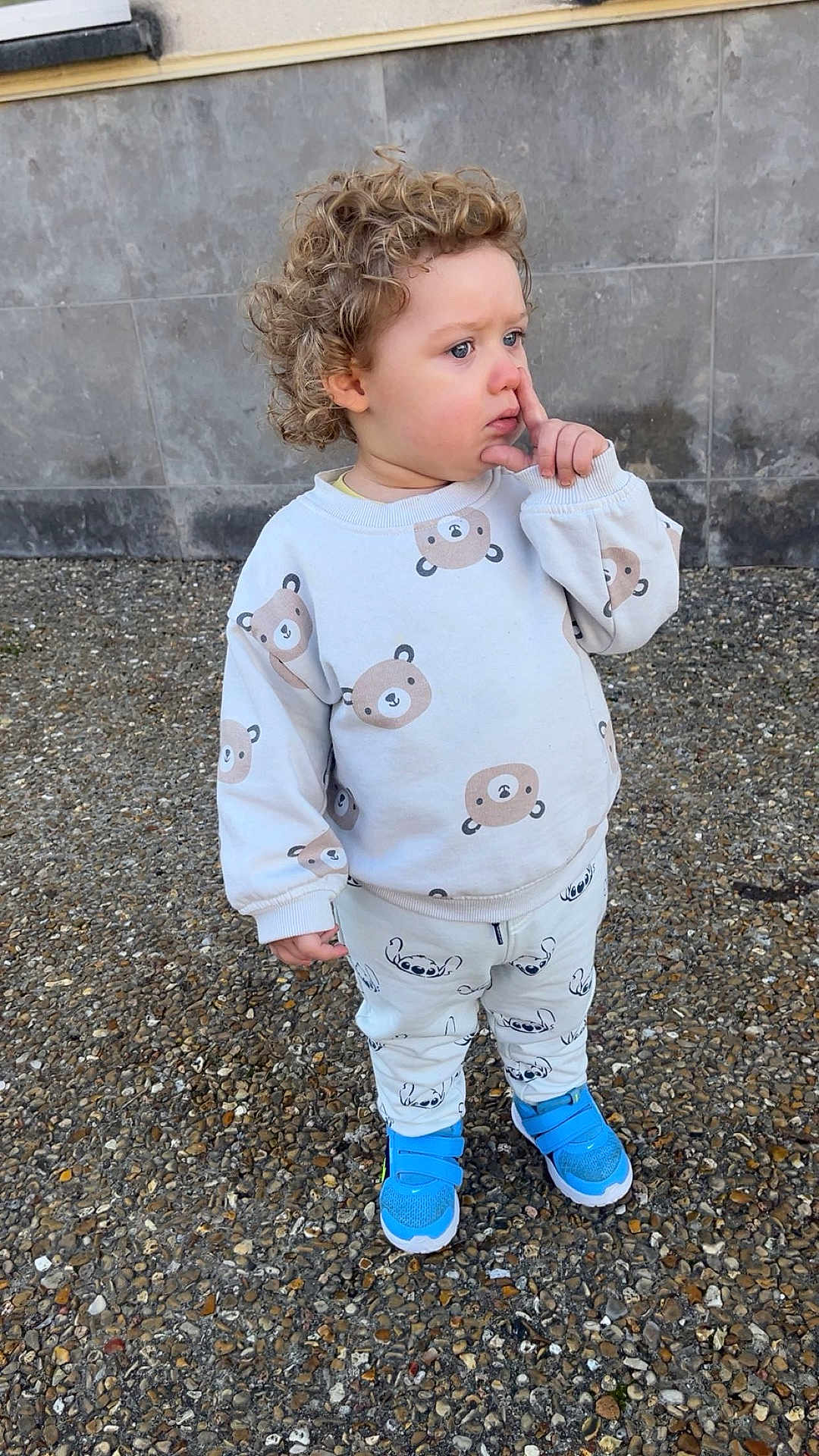 Maylon participe au concours pour gagner de l'argent avec cette photo : child, toddler, curly_hair, sweater, bear_print, patterned_pants, blue_sneakers, standing, gravel_ground, stone_wall, outdoor, hand_to_face, picking_nose, thoughtful_expression, portrait, casual_clothing, single_face, shoes, daytime, cute