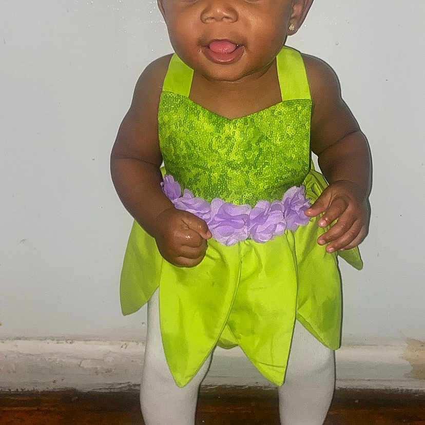 Jazlynn is registered to the contest to win money with this photo: baby, child, cute, earrings, girl, green_dress, happy, headband, indoors, person, pink_socks, plain_wall, playful, portrait, purple_flowers, smiling, standing, toddler, white_tights, wooden_floor