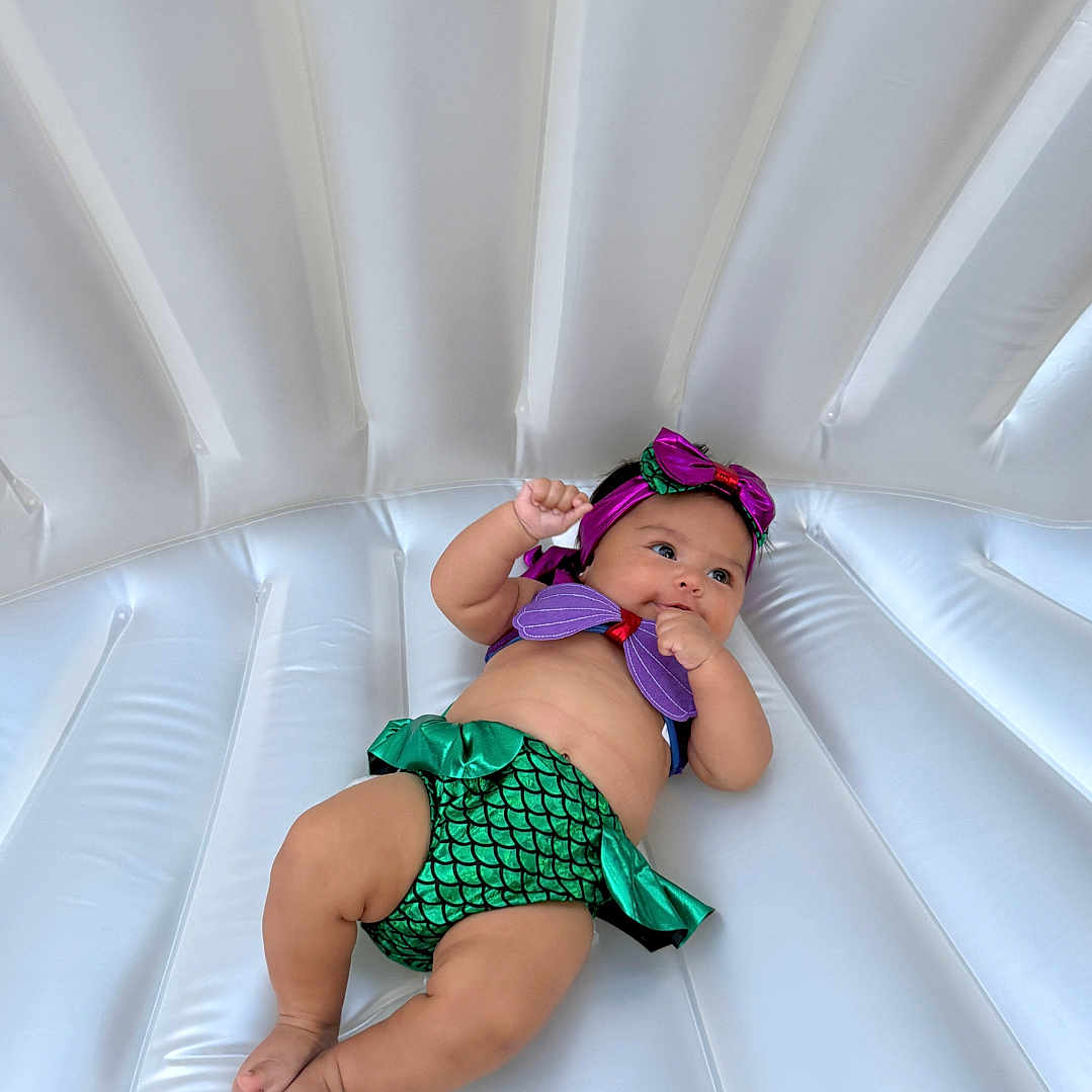 Serena is registered to the contest to win money with this photo: baby, infant, mermaid_costume, headband, purple, green, inflatable, white, cute, child, lying_down, playful, soft_surface, portrait, indoors, clothing, small_person, baby_girl, colorful, relaxed