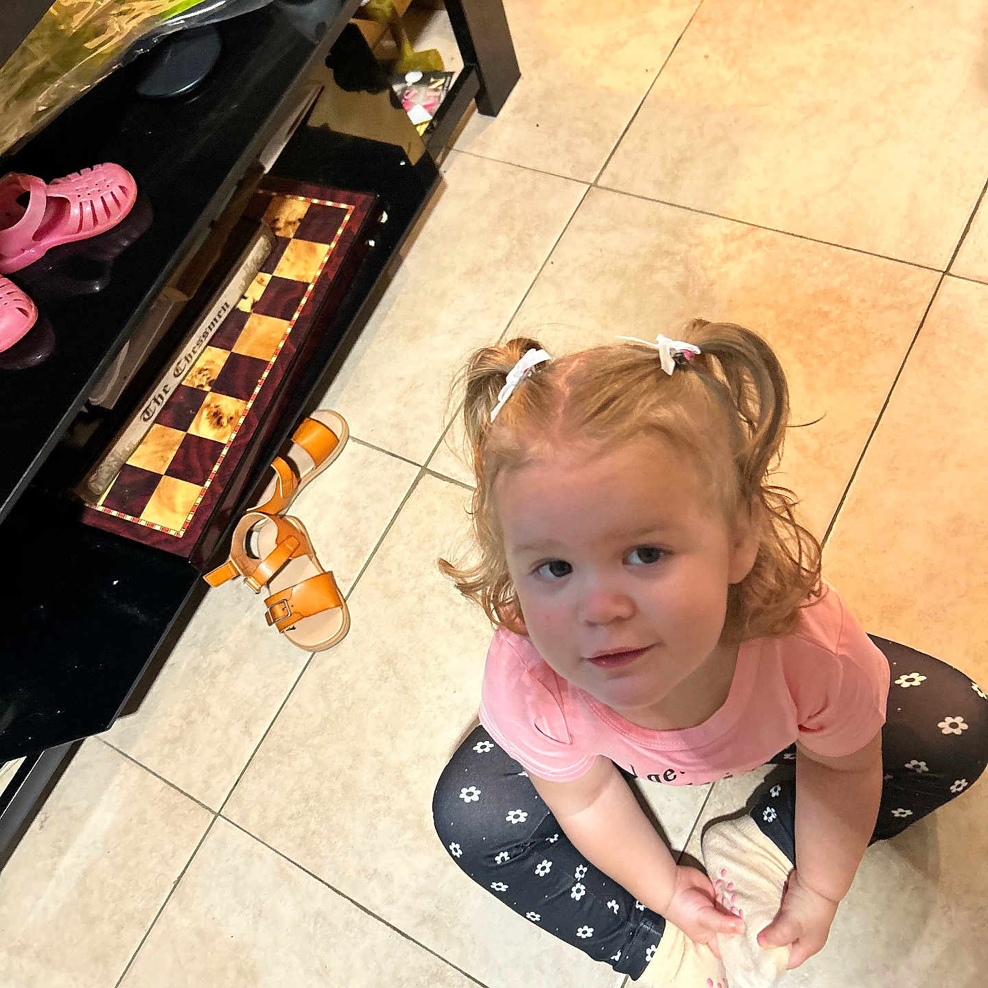 Charli joined the competition — help win amazing prizes! black_leggings, casual, chessboard, child, curly_hair, girl, home, indoor, looking_up, orange_sandals, pigtails, pink_shirt, pink_shoes, shelf, sitting, smile, socks, tile_floor, toddler, white_flowers
