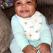 Kenyan joined the competition — help win amazing prizes! baby, bib, blanket, bright_eyes, chubby_cheeks, closeup, couch, cozy, dimple, feet, hands, infant, living_room, onesie, patterned_fabric, portrait, sitting, smiling, teeth, toes