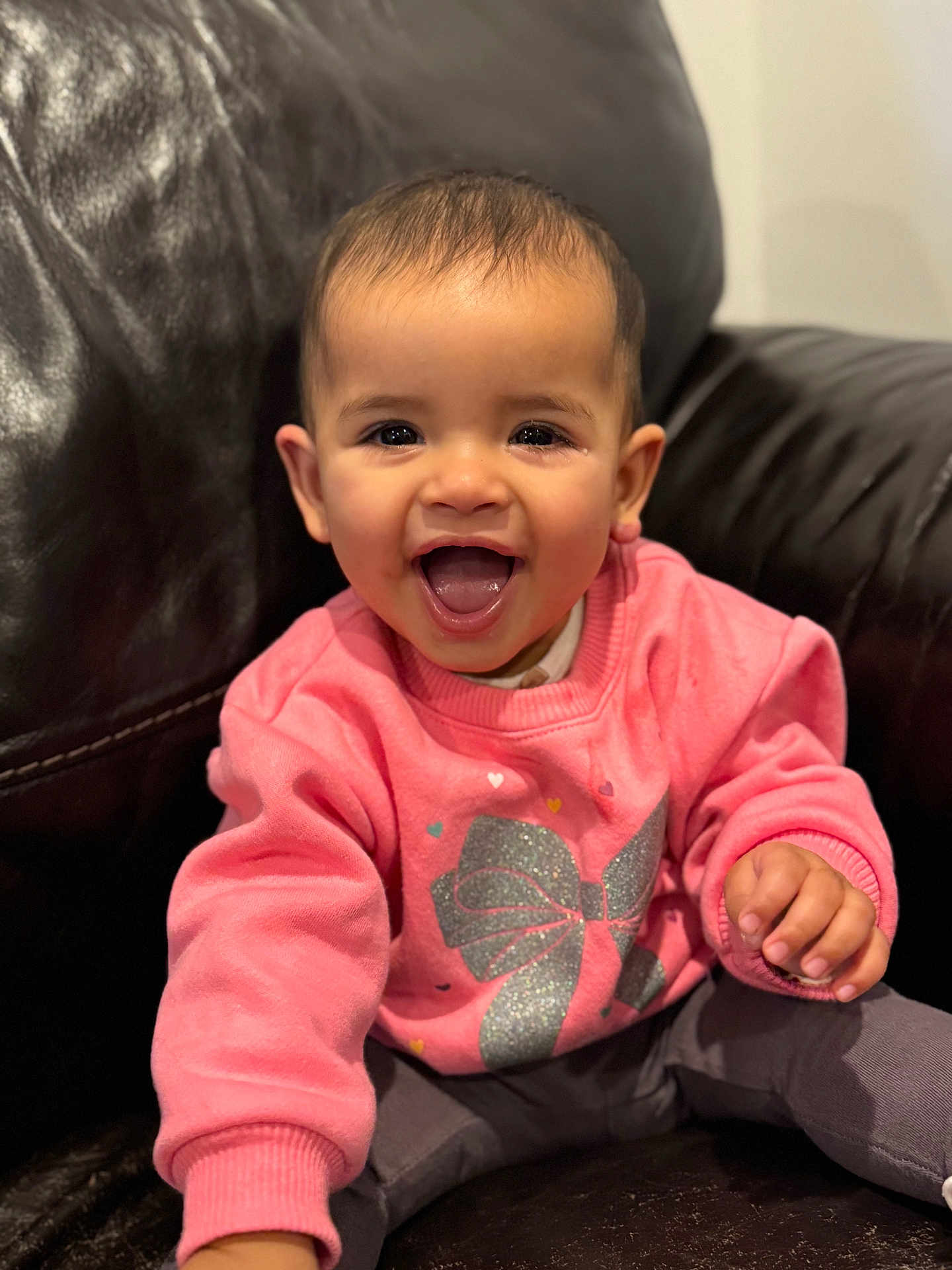 Ayla joined the competition — help win amazing prizes! baby, child, smiling, happy, face, pink_sweater, glitter, bow, sitting, leather_couch, indoor, portrait, cute, joyful, young_child, clothing, person, hand, head, eyes