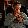 woman, kitten, cat, indoor, chair, casual_clothing, playing, pet, light, sunglasses, sitting, hand, holding, home, shelf, furniture, gentle, interaction, gray_sweater, intimate