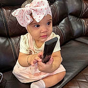Emily joined the competition — help win amazing prizes! baby, bow, bracelet, child, clothing, couch, curious, cute, furniture, headband, indoors, leather, person, pink, remote_control, sitting, small_hands, socks, toddler, white