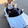 toddler, child, toy_car, sunglasses, white_tank_top, shorts, sidewalk, outdoor, sunlight, small_bottle, miniature_vehicle, summer, cute, fashion, playtime, casual_clothing, happy, young_child, driving, fun