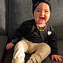baby, child, smiling, laughing, sitting, black_cardigan, headband, bow, leopard_print_socks, cream_pants, couch, gray_couch, indoor, happy, cute, jewelry, necklace, heart_patch, shadow, sunlight