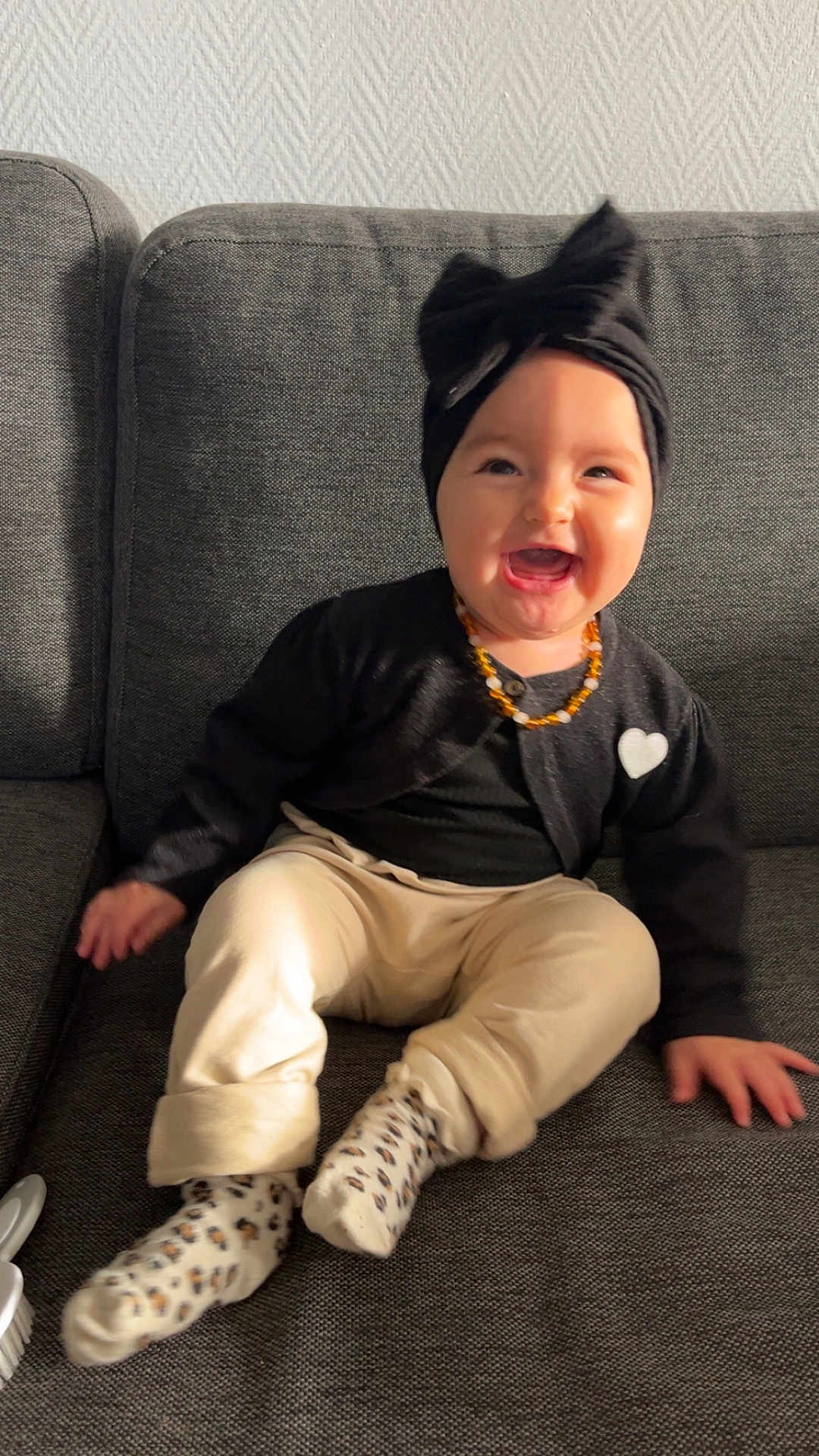 Valentina participe au concours pour gagner de l'argent avec cette photo : baby, child, smiling, laughing, sitting, black_cardigan, headband, bow, leopard_print_socks, cream_pants, couch, gray_couch, indoor, happy, cute, jewelry, necklace, heart_patch, shadow, sunlight
