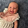 Millie is registered to the contest to win money with this photo: baby, smiling, infant, pink_bib, white_dress, carpet, happy, cute, portrait, child, face, skin, hand, lying_down, indoors, closeup, young, person, apparel, textile
