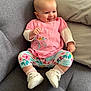 baby, smiling, pink_shirt, floral_pants, white_shoes, socks, couch, gray_fabric, indoor, child, happy, cute, infant, seated, comfortable, portrait, young_child, casual_clothing, home, cozy